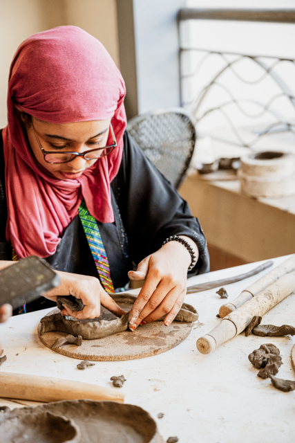 A participant in a pink headscarf shapes clay on a pottery work board.