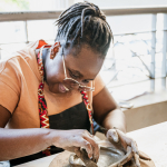 Woman in glasses and an apron shapes a clay dish by hand.