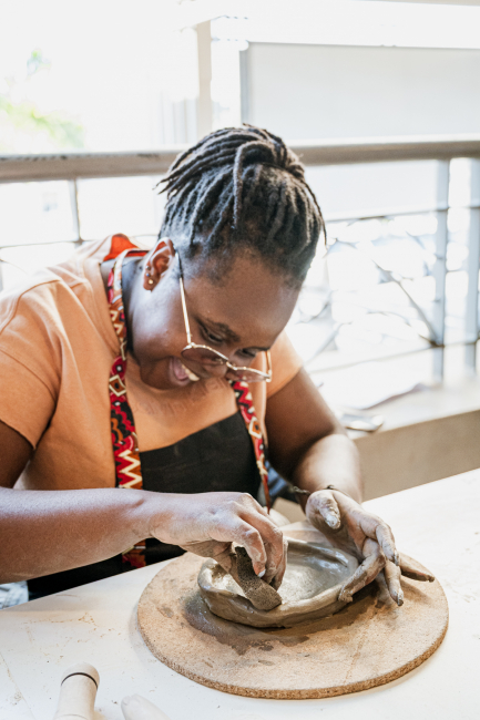 Woman in glasses and an apron shapes a clay dish by hand.