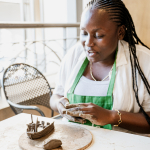 Woman in a green apron works with clay beside an unfinished pottery piece.