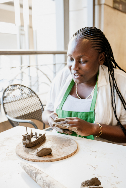 Woman in a green apron works with clay beside an unfinished pottery piece.