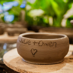 A handmade clay bowl with a handwritten message and heart engraving rests on a wooden board.