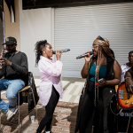 Two singers perform together with microphones while musicians accompany them.