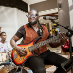Bassist playing an electric bass with other musicians visible behind him