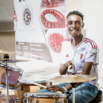 Smiling drummer seated at a drum kit with a Coming Soon poster visible in the background.