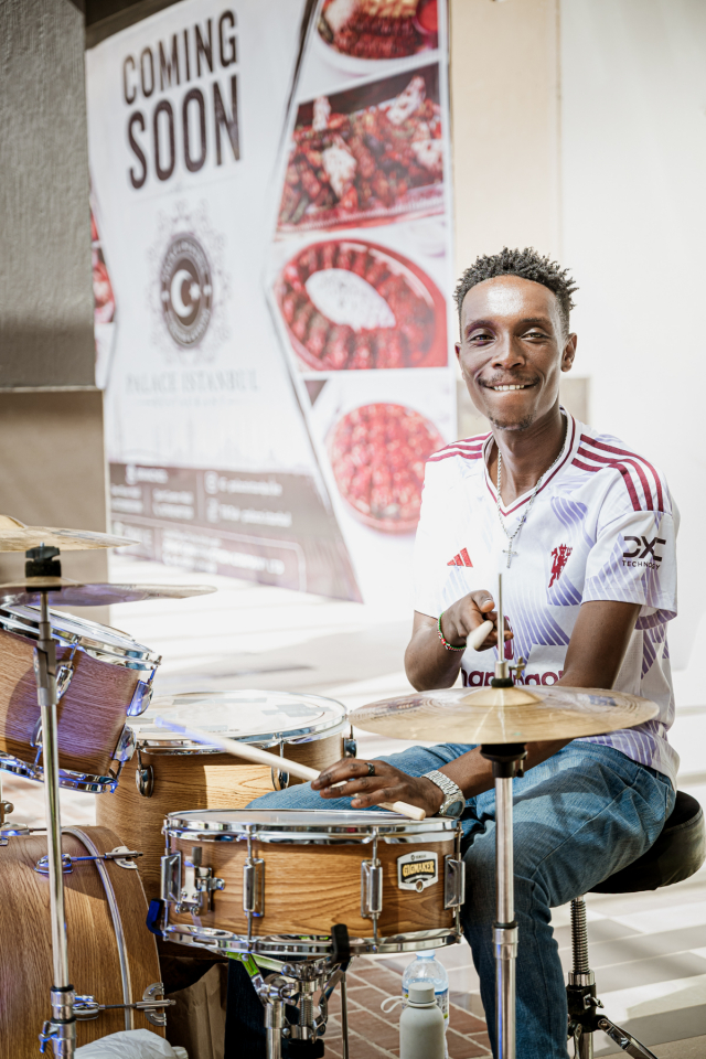 Smiling drummer seated at a drum kit with a Coming Soon poster visible in the background.