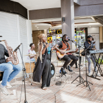 A five-member band performs together in an open mall area.