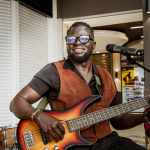 Smiling bassist holding a sunburst electric bass beside a microphone.