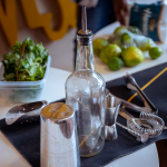 A cocktail preparation station with a pour bottle, shaker, jigger, herbs, limes, and bar tools.