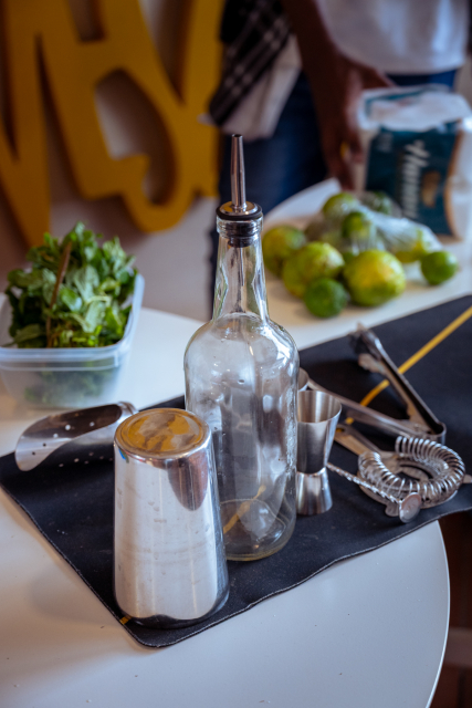 A cocktail preparation station with a pour bottle, shaker, jigger, herbs, limes, and bar tools.