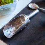 A metal cocktail strainer and spoon rest on a black bar mat beside fresh herbs.