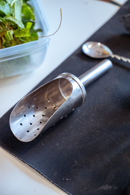 A metal cocktail strainer and spoon rest on a black bar mat beside fresh herbs.