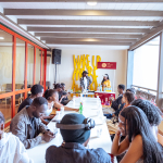 A large group sits around a long table facing a presentation area with WAKE UP lettering on the wall.