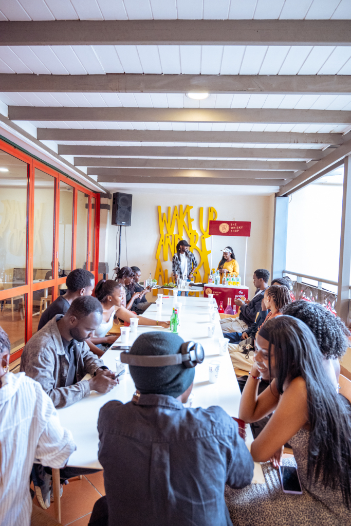 A large group sits around a long table facing a presentation area with WAKE UP lettering on the wall.