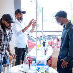 Three people interact beside a tasting counter as one man gestures and another holds a cup.