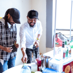 Two men smile while preparing samples among bottles, herbs and cups on a tasting table.