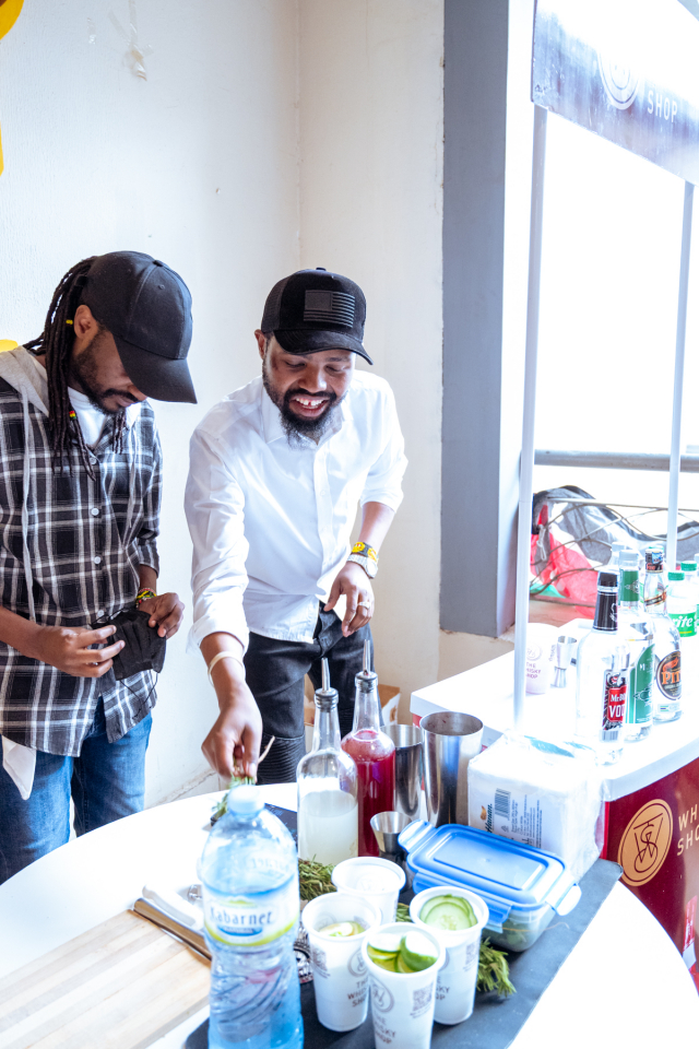 Two men smile while preparing samples among bottles, herbs and cups on a tasting table.
