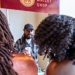 The Whisky Shop representative holds a tasting cup behind a bottle table while attendees stand in the foreground