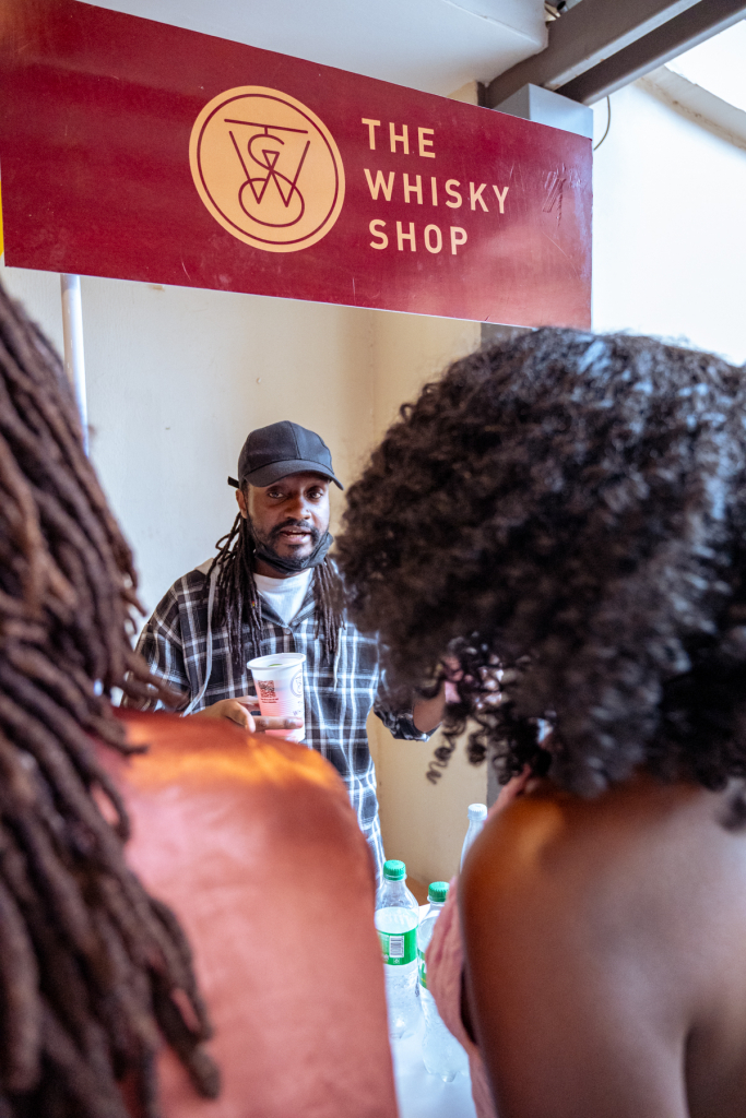 The Whisky Shop representative holds a tasting cup behind a bottle table while attendees stand in the foreground