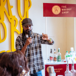 Speaker holds a microphone beside a The Whisky Shop sign and a display of bottles