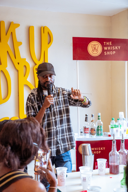 Speaker holds a microphone beside a The Whisky Shop sign and a display of bottles