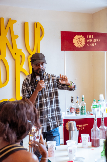Speaker holds a microphone beside a The Whisky Shop sign and a display of bottles