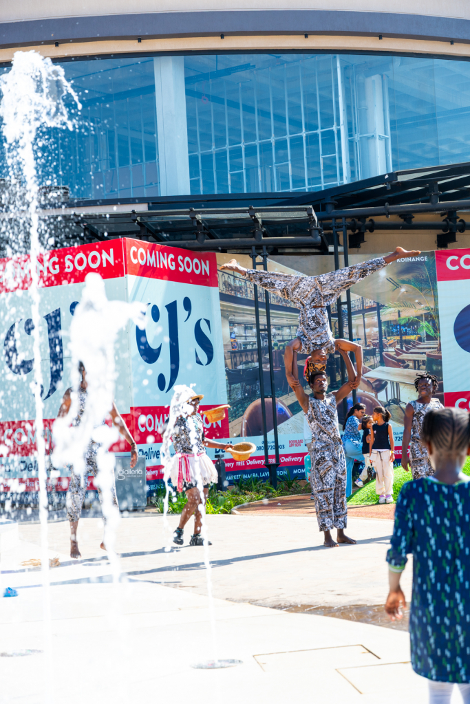 A performer holds an elevated pose behind a fountain in the mall plaza.