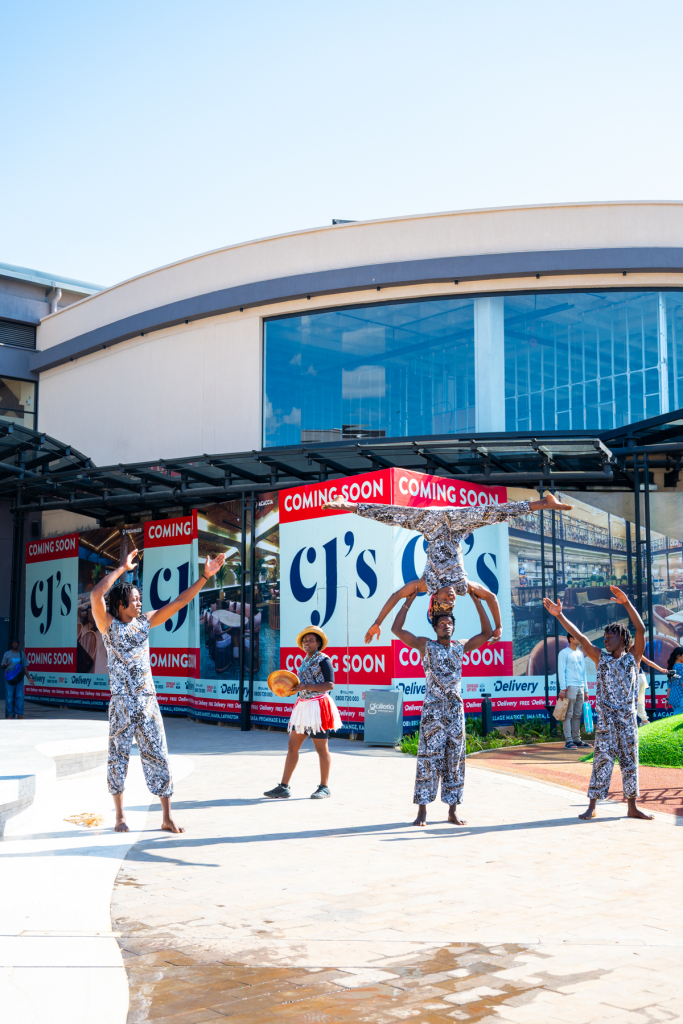 Performers raise their arms in formation outside a shopping mall entrance.