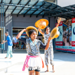 DZ6 1229 A performer holds a woven hat above her head while another performer displays hats behind her outside the mall.