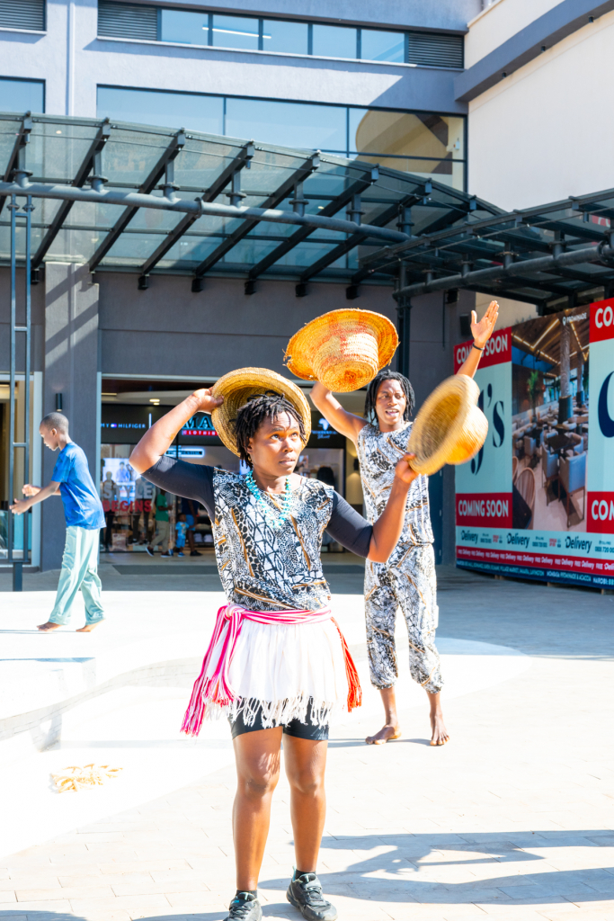 A performer holds a woven hat above her head while another performer displays hats behind her outside the mall.