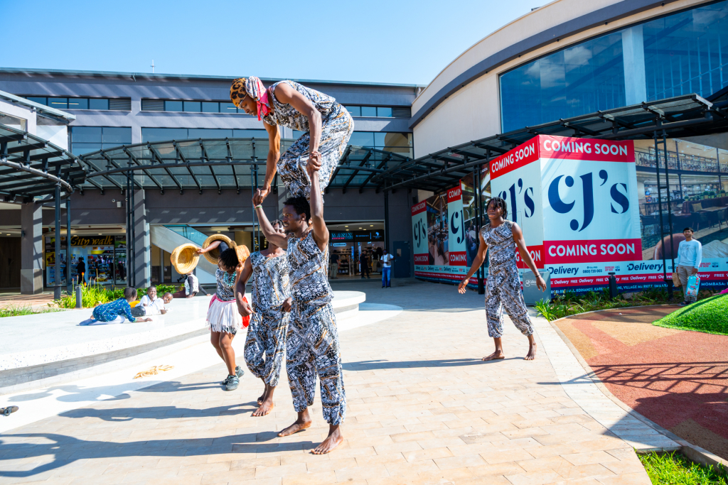 A group of performers presents an acrobatic routine in the mall courtyard with storefronts behind them.