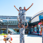 DZ6 1263 Performers in matching patterned outfits form a human acrobatic pose outside the shopping mall.
