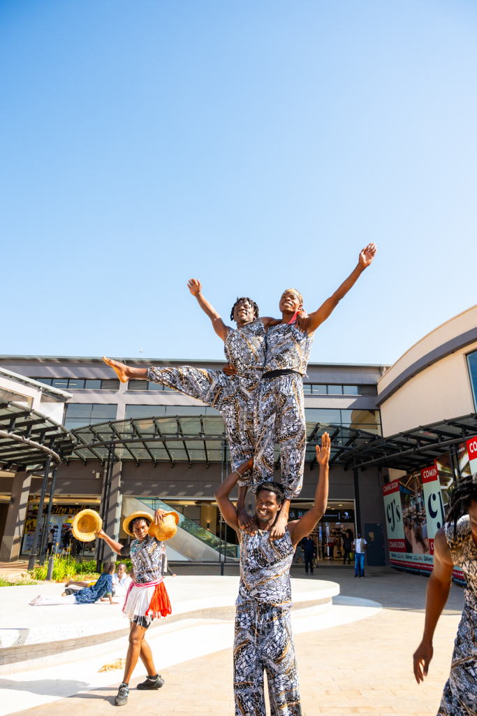 Performers create a tall human pyramid outside the shopping mall while other performers stand nearby.