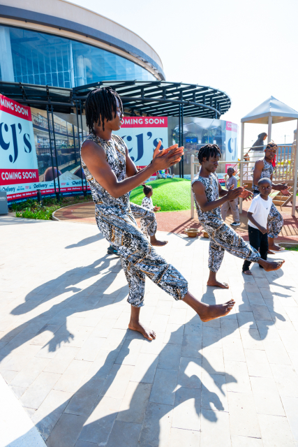 A dancer performs an extended leg movement while other performers and children watch nearby.