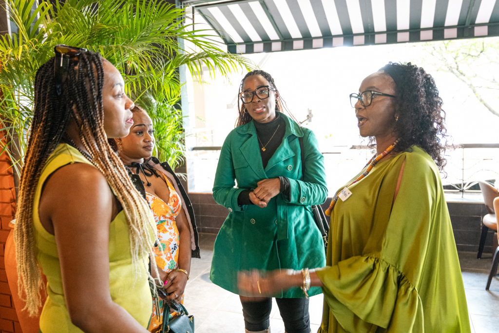 Four women stand together in conversation near plants and bright windows.