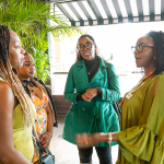 DZ6 1482 Four women stand together in conversation near plants and bright windows.