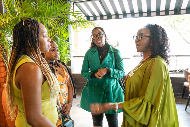 Four women stand together in conversation near plants and bright windows.