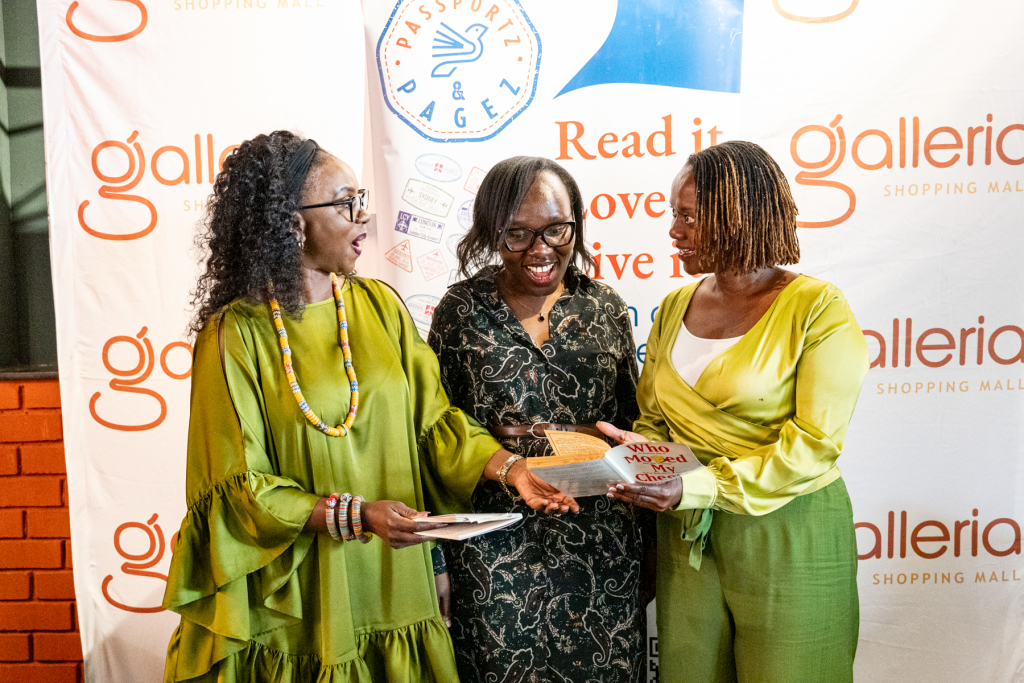 Three women smile and share a booklet or card in front of branded event banners.