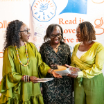 DZ6 1558 Three women smile and share a booklet or card in front of branded event banners.
