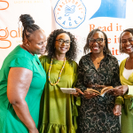 DZ6 1588 Four women smile together while looking at an open book in front of event branding.