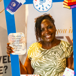 DZ6 1707 Smiling woman holds a blue reading-themed photo frame decorated with books, passports, and event graphics.