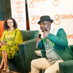DZ6 2254 A man wearing a hat speaks into a microphone while seated beside a listening woman at a mall event.