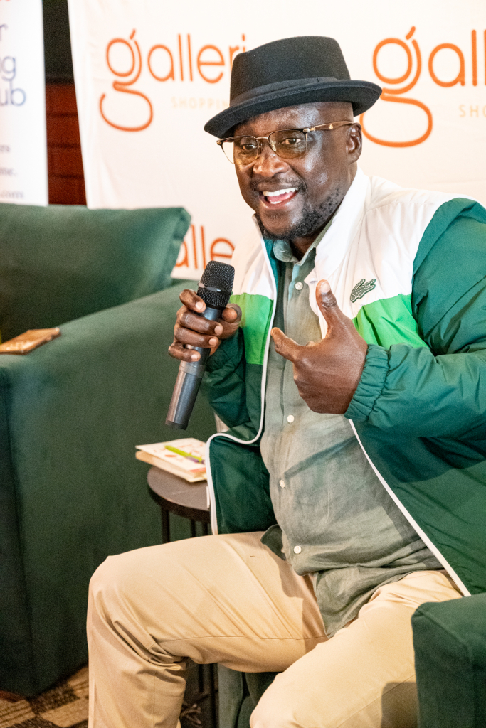 A smiling man in a hat gestures while holding a microphone in a lounge chair.