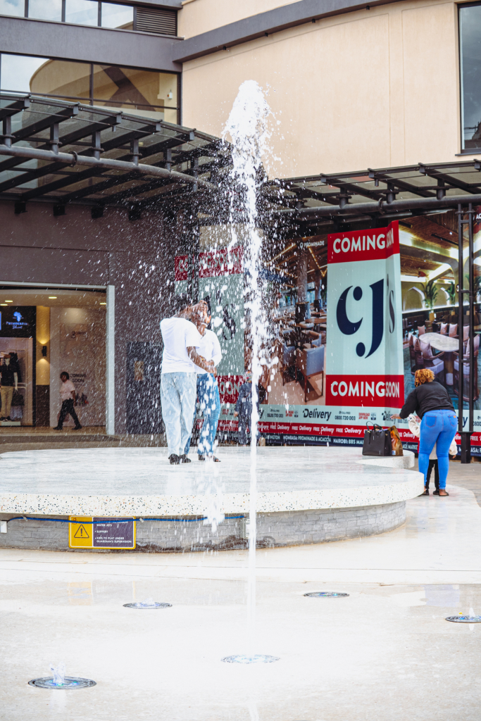A fountain sprays in front of mall storefronts and people gathered in the courtyard